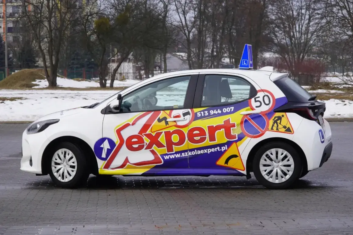 Biały samochód szkoleniowy z napisem "Sexpert" i znakami drogowymi, gotowy do prowadzenia pojazdu.