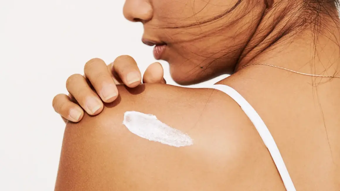 Eine Hand trägt eine weiße Creme auf die Schulter auf. Die Creme könnte Panthenol oder Bepanthen enthalten, um die Haut zu pflegen.