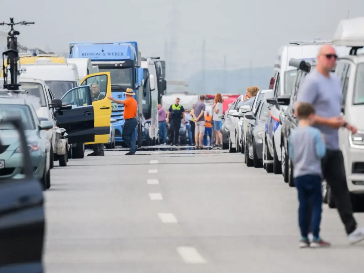 Stau auf der Autobahn. Ein Mann in orangefarbener Jacke und Hut steht neben einem gelb-schwarzen Transporter, dessen Tür offen ist. Viele Autos und LKW stehen still, als ob sie trotz Fahrverbot unterwegs wären.