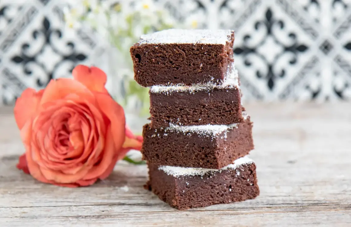 Schokoladige Brownies mit Datteln, bestäubt mit Puderzucker, neben einer zarten orangefarbenen Rose auf Holztisch.