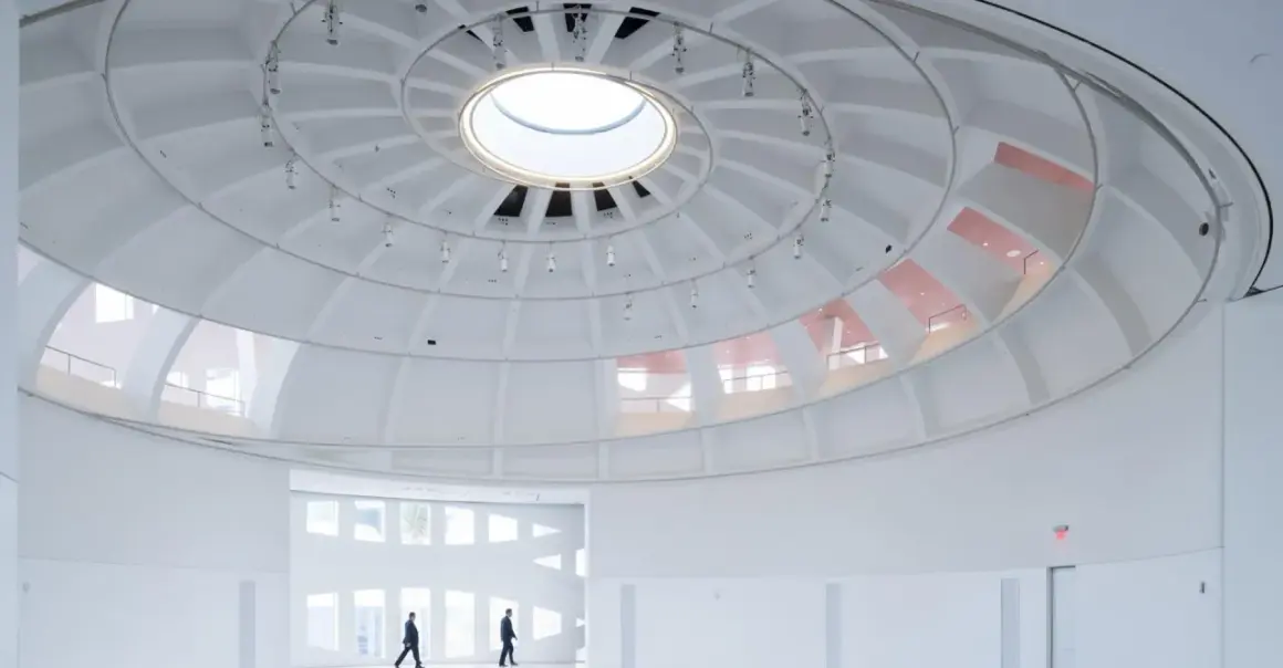 The expansive, white interior of the Faena Art Project Room features a striking domed ceiling with a central skylight. Two figures walk through the minimalist space.