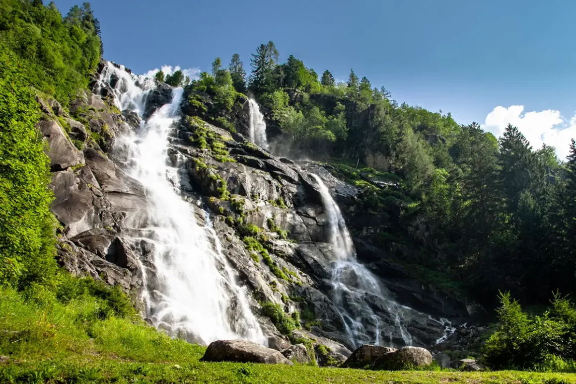 Cascate maestose nel Val di Genova, con acqua che scende impetuosa su rocce scure, circondate da fitta vegetazione verde e alberi imponenti sotto un cielo azzurro.
