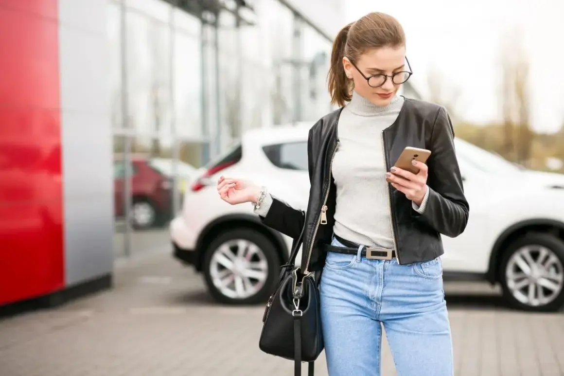 Kobieta w okularach i skórzanej kurtce sprawdza telefon, stojąc obok białego auta. Zastanawia się, kto może wziąć auto w leasing.