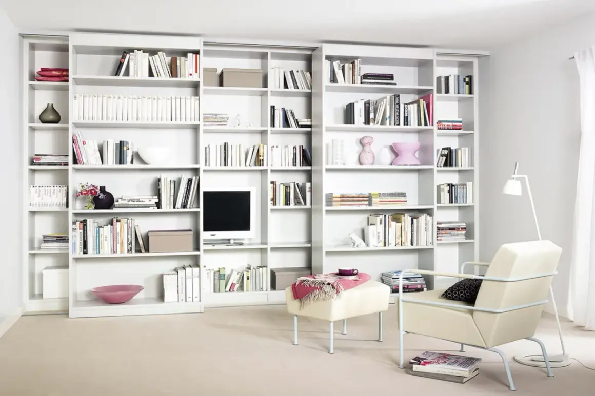Eine moderne, weiße **eigene Bibliothek** mit vielen Büchern, Dekorationen und einem Fernseher. Ein Sessel und ein Hocker laden zum Verweilen ein.