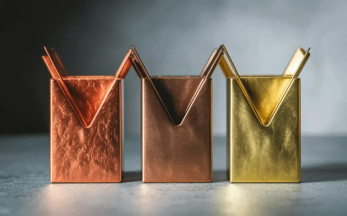 Trois porte-stylos aux formes géométriques, en laiton et cuivre, aux textures variées.