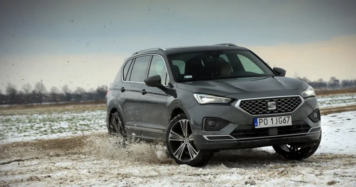Szary Seat Tarraco SUV przemierza błotniste, zaśnieżone pole, wzbijając chmury brudu.