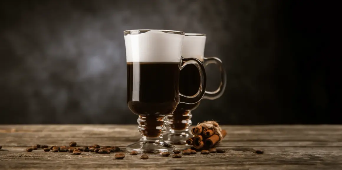 Två glas med kaffe med glass och vispgrädde, omgivna av kaffebönor och kanelstänger på ett träbord.