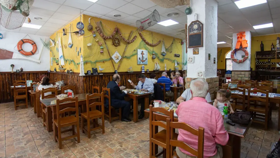 Gente comiendo en la abacería el arrecife, decorada con motivos marineros. Mesas de madera y paredes amarillas con redes y barcos.