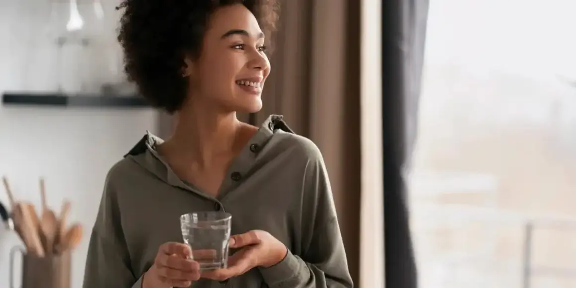 Une femme souriante tient un verre d'eau, un geste simple qui symbolise le luxe de l'eau de riche, une hydratation pure et précieuse.