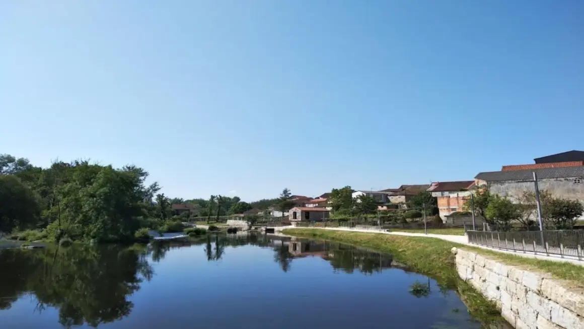 Tranquila playa fluvial en Leiro, con casas tradicionales reflejándose en el agua serena bajo un cielo azul.