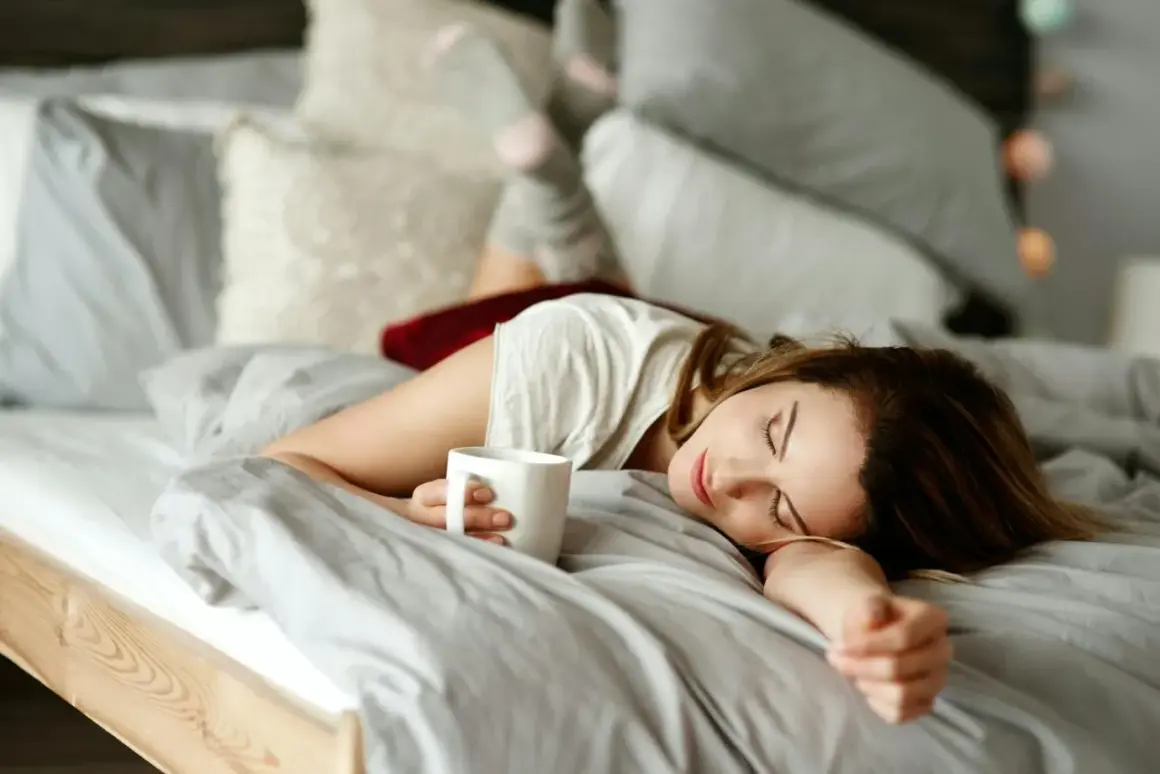 En kvinna sover i sängen med en kaffekopp i handen. Kaffe bra eller dåligt? Kanske en tupplur är bättre.
