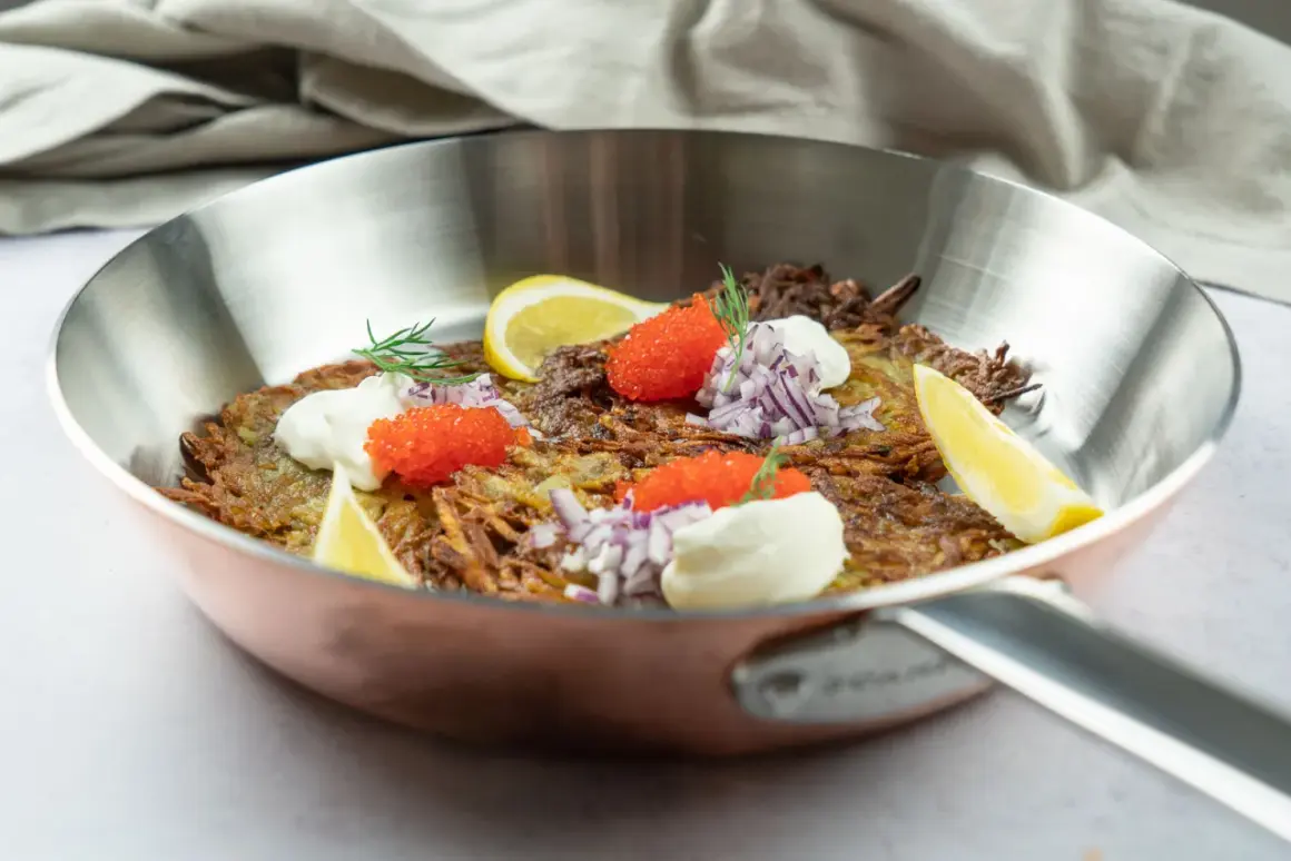 En gyllene råraka i stekpanna, toppad med gräddfil, rödlök, rom och citronklyfta. Ett klassiskt råraka recept som ser utsökt ut.