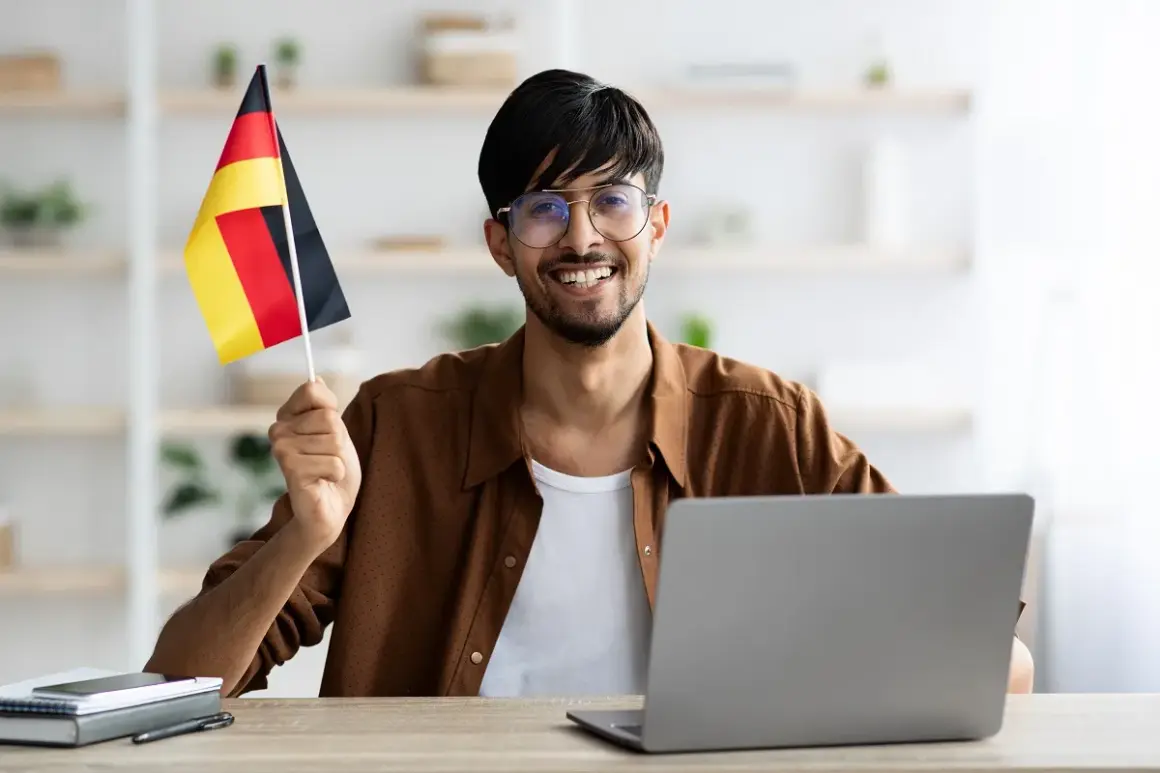 Uśmiechnięty mężczyzna z flagą Niemiec przy komputerze, planuje otwarcie konta w niemczech bez meldunku.