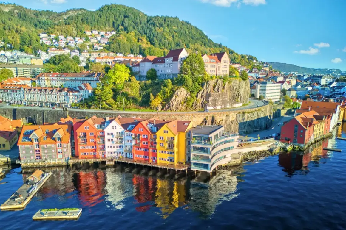 Kolorowe domy nad wodą w Bergen, idealne na wycieczki do Norwegii. W tle zielone wzgórza i zabudowania.