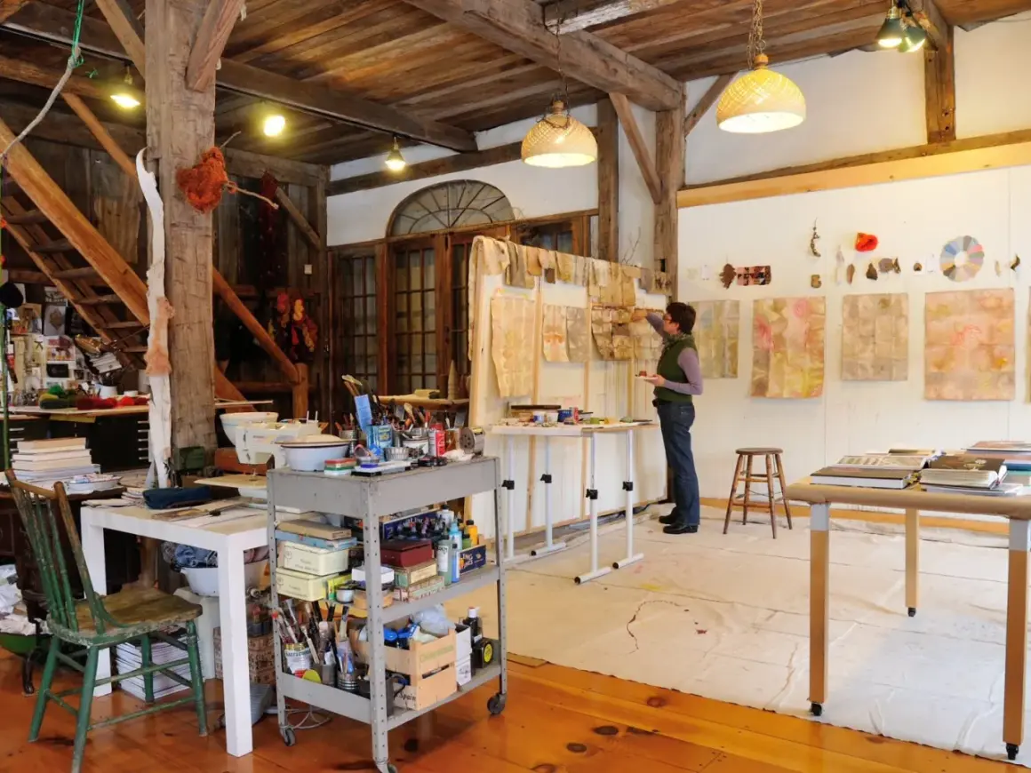 A woman works in a rustic studio, surrounded by art supplies and botanical prints, reminiscent of famous artists studios.
