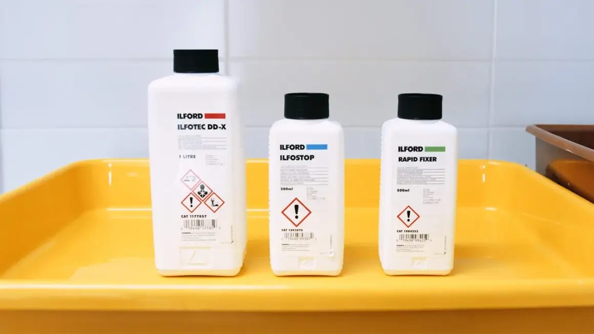 Three Ilford bottles for developing, stopping, and fixing film sit in a yellow tray, ready for creating the best black and white film.