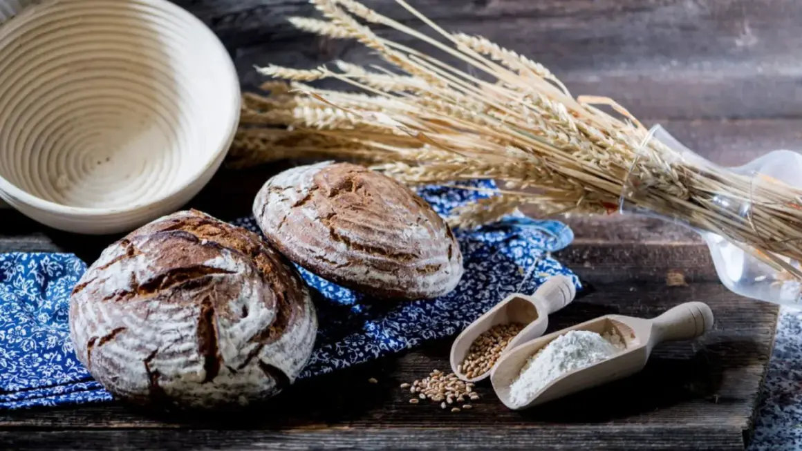 Zwei rustikale Brote, Weizenähren und Holzschaufeln mit Getreide und Mehl. Eine Bäckerei-Szene, die die Vielfalt einer **Getreide Sorte** zeigt.