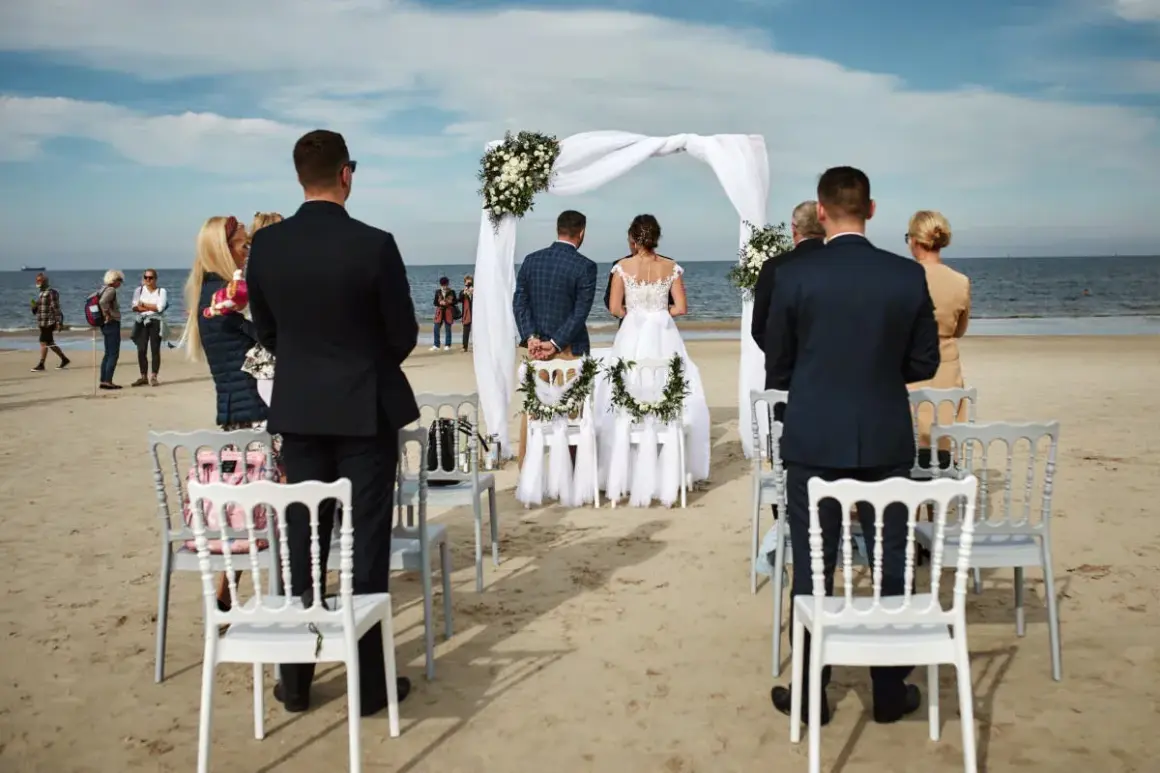 Piękny ślub na plaży. Para młoda stoi przed białym baldachimem, goście obserwują ceremonię.
