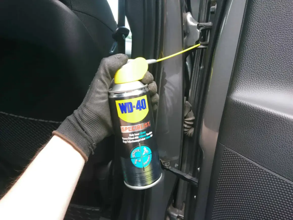 Ręka w rękawiczce aplikuje biały smar litowy WD-40 na zawiasy drzwi, rozwiązując problem czym smarować zawiasy w drzwiach.