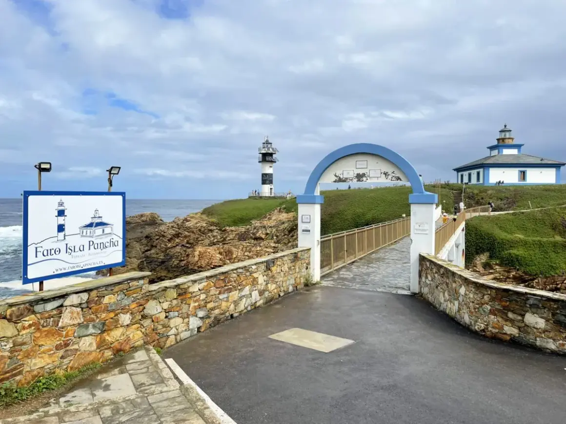 El faro de Ribadeo, con su icónico diseño blanco y negro, se alza sobre un acantilado verde. Un puente arqueado conduce a la isla.