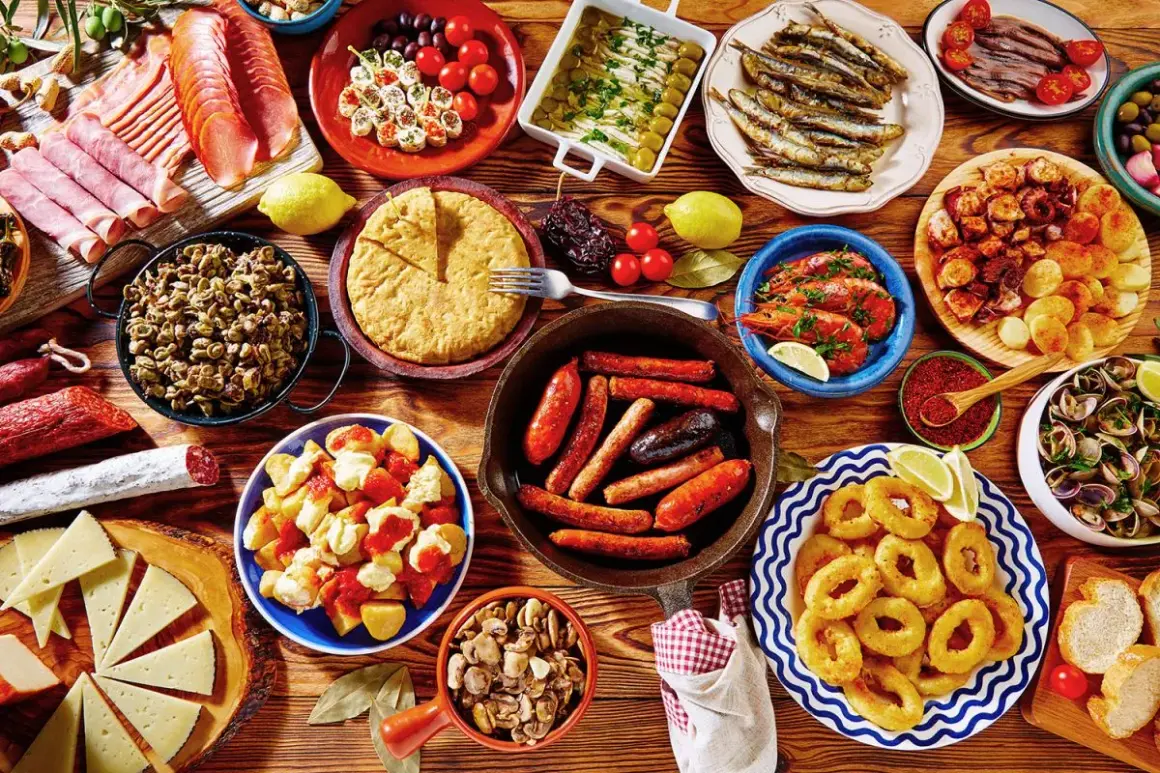Obfity stół pełen hiszpańskich tapas: kiełbaski, krewetki, kalmary, owoce morza, ser, wędliny i tortilla.