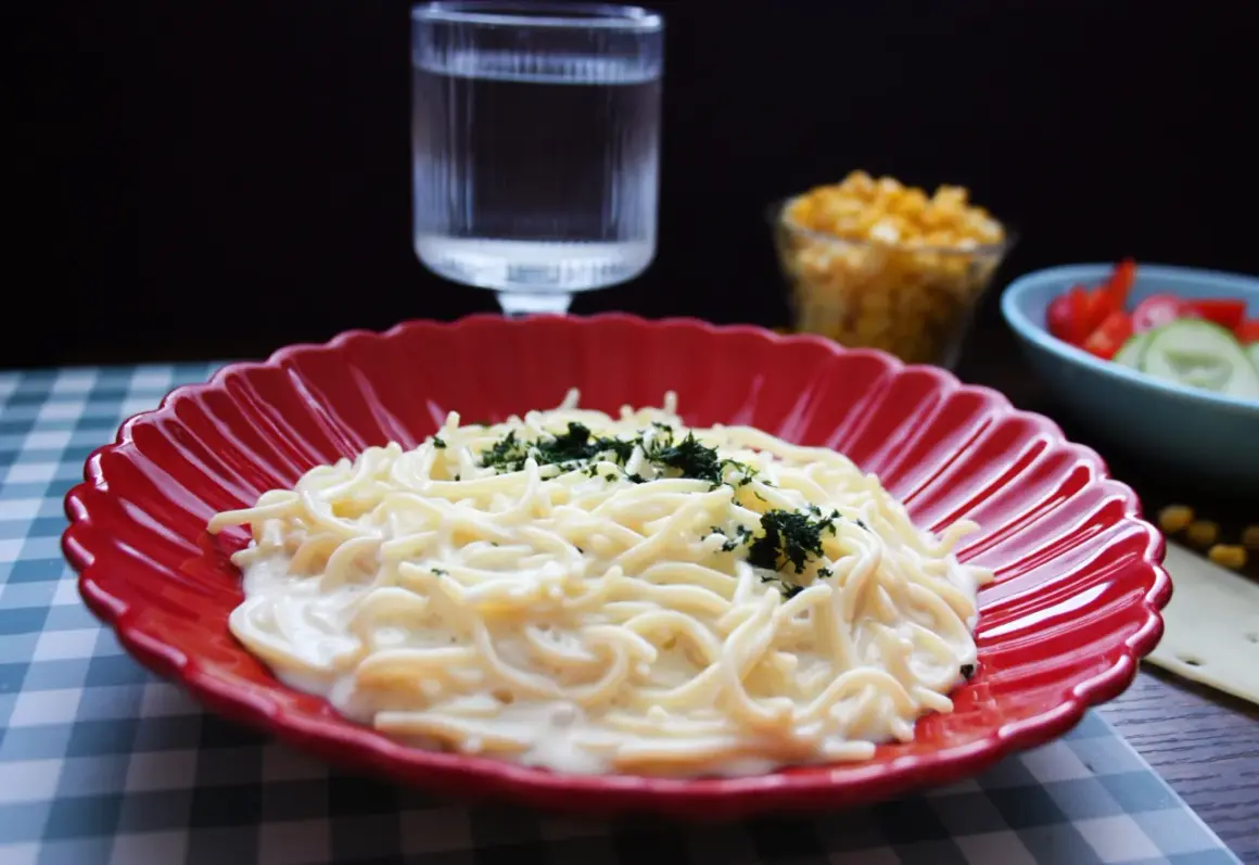 En krämig mac and cheese med persilja på en röd tallrik. Bredvid syns en sallad och ett glas vatten.