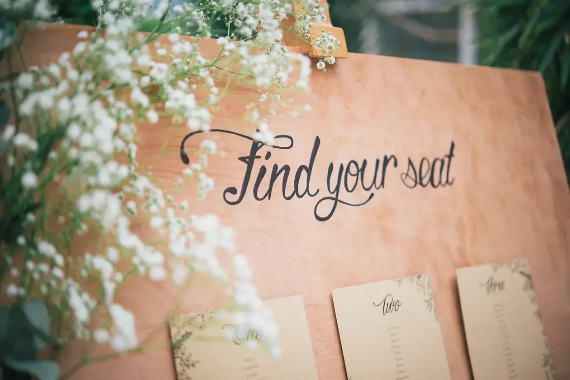 Panneau en bois avec l'écriture "Find your seat" et des fleurs. Des cartes de placement sont fixées en dessous.
