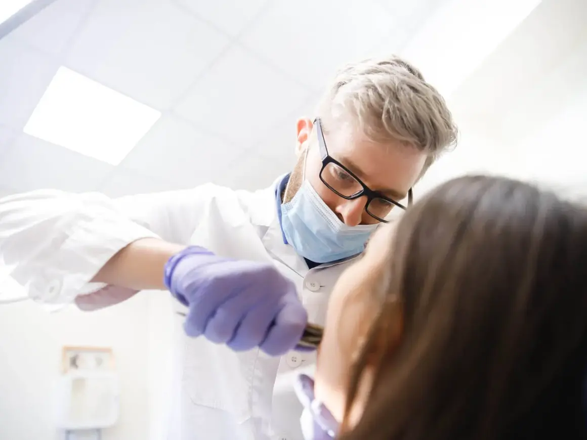 Dentysta w masce i rękawiczkach wykonuje borowanie zęba pacjentowi.