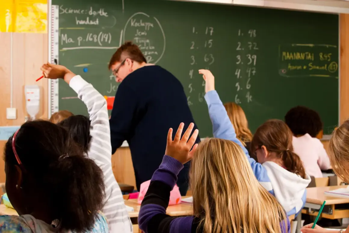 Schüler heben in einer finnischen Schule die Hände. Eine Lehrerin steht vor einer Tafel mit mathematischen Aufgaben.