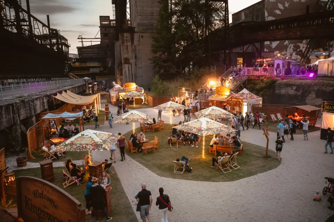 Besucher genießen das Ostrava Festival in einer ehemaligen Industrieanlage. Stände, Sitzbereiche und Lichter schaffen eine festliche Atmosphäre.