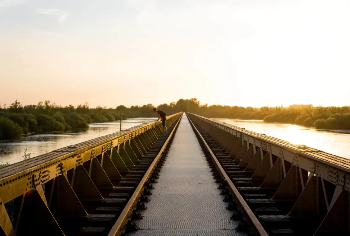 Een man leunt over een spoorbrug bij zonsondergang. De gouden gloed verlicht de rivier en de weelderige vegetatie, wat zorgt voor mooie foto locaties.