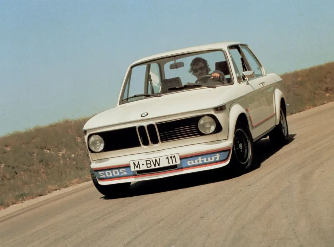 Białe BMW E20 z kolorowymi pasami na zderzaku i czerwonymi akcentami na karoserii jedzie po krętej drodze.