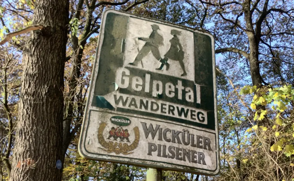 Schild am wanderparkplatz Gelpetal weist den Weg. Silhouetten von Wanderern und Bierwerbung.