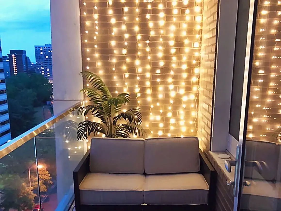 Przytulny balkon w bloku z sofą i palmą, ozdobiony ciepłym oświetleniem.