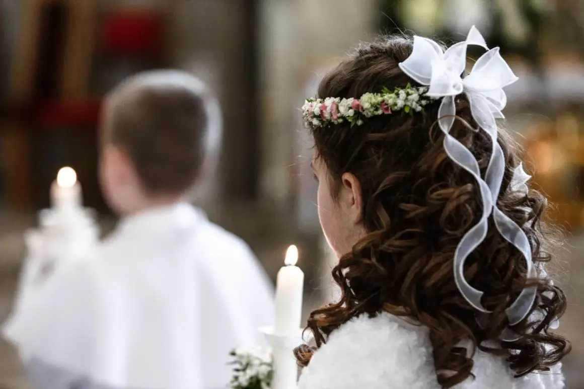 Dziewczynka z wiankiem i kokardą na głowie, trzymająca świecę. W tle chłopiec w białej komży. Biały tydzień po komunii.