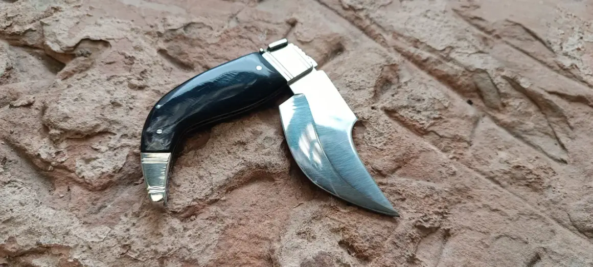 Navaja capaora de mango negro y hoja curva, lista para la acción sobre una superficie rocosa.