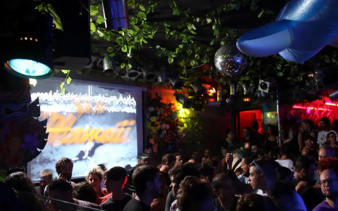 Folla in un locale notturno a Barcellona, con luci stroboscopiche, piante finte e un grande squalo gonfiabile. Atmosfera da festa al Moog.
