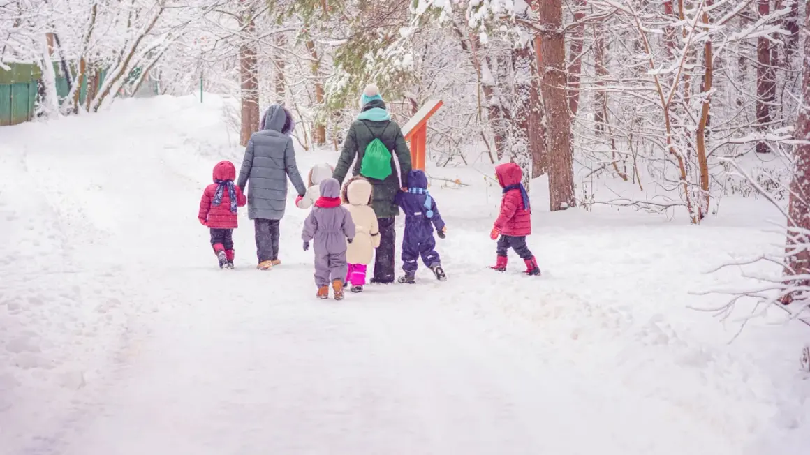 Schneebedeckter Weg im Wald. Eine Gruppe Kinder und eine Erzieherin gehen zum Kindergarten.