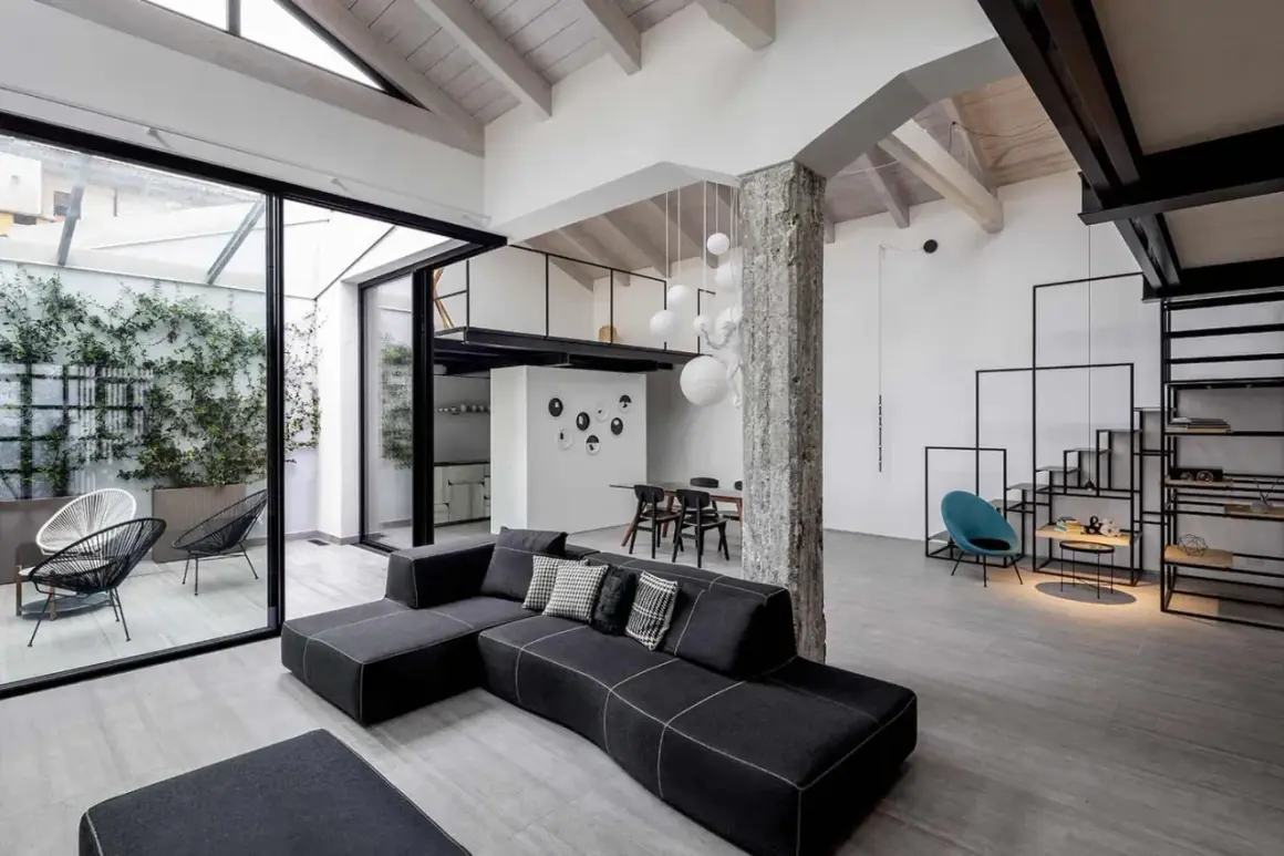 Un ampio soggiorno con tetto a vista e un soppalco, che mostra cos'è un loft: spazio aperto, design industriale e un tocco di verde da un patio interno.