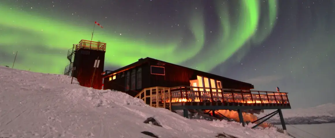 En snöig sluttning leder upp till en mysig aurora sky station. Norrskenet dansar grönt över stjärnorna.