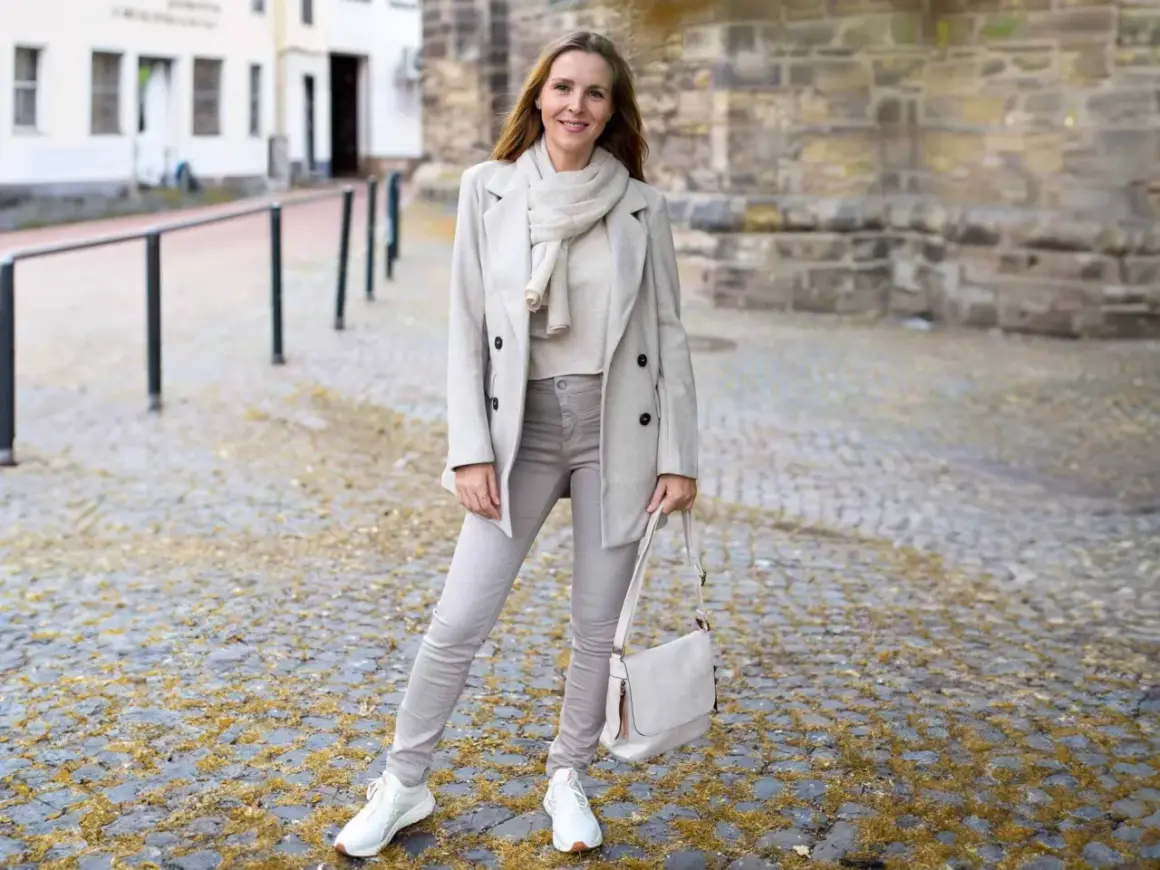 Frau in beige grau farbe Outfit mit Mantel, Schal, Hose und Tasche steht auf Kopfsteinpflaster.