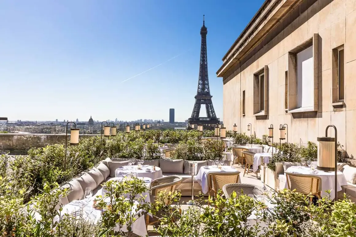 Terrasse d'un restaurant chic avec vue sur la Tour Eiffel. Tables dressées, végétation luxuriante et lanternes créent une ambiance magique.