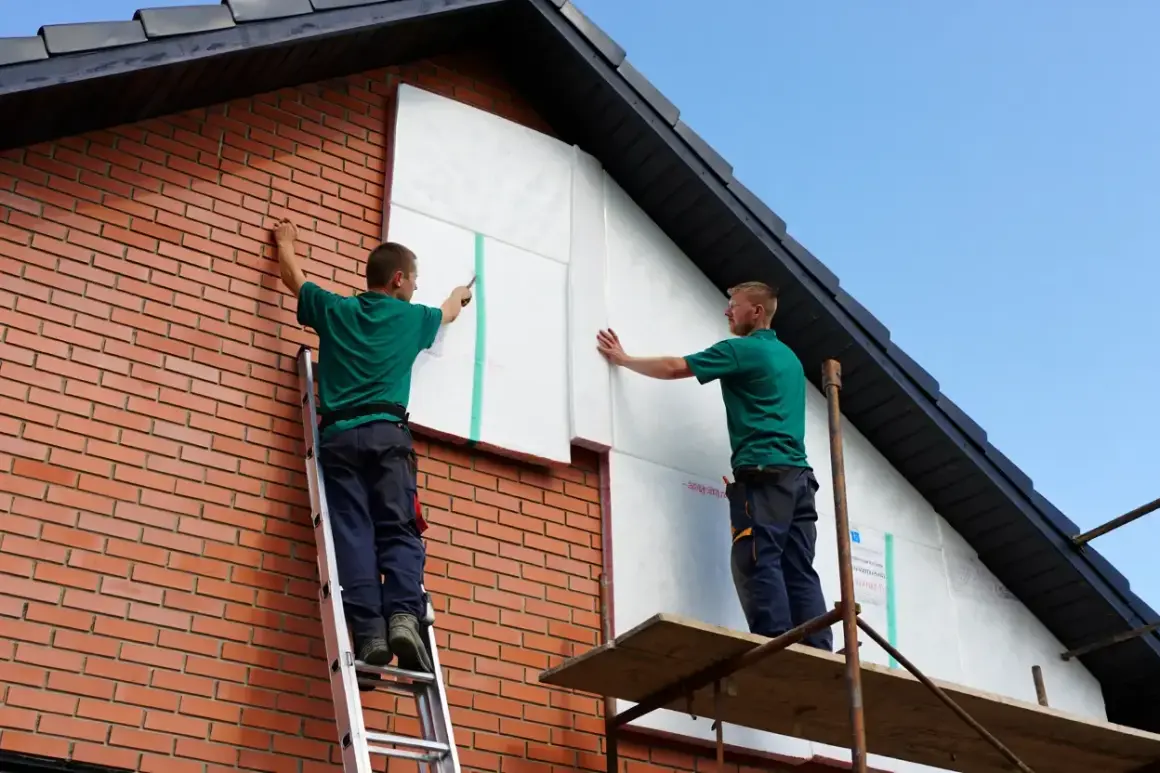 Deux ouvriers réalisent l'isolation par l'extérieur soi même d'une maison en briques, fixant des panneaux isolants blancs sur le pignon.