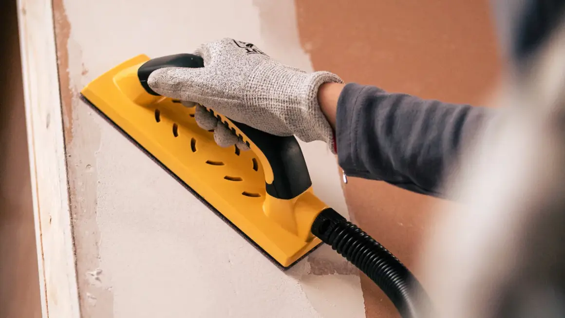 Mano enguantada usando una lijadora amarilla para lijar paredes, preparando la superficie para pintar.