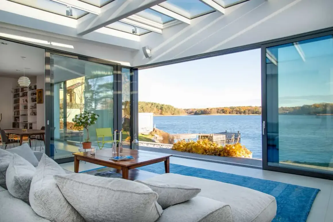 Soggiorno luminoso con vista lago, divano grigio e tavolino in legno. L'ampia vetrata e il soffitto trasparente creano un perfetto stile coastal.