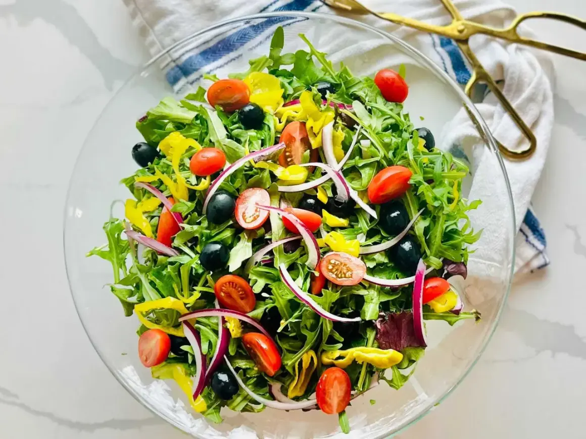 En fräsch italiensk sallad i en glasskål, fylld med gröna blad, körsbärstomater, svarta oliver, rödlök och gul paprika.
