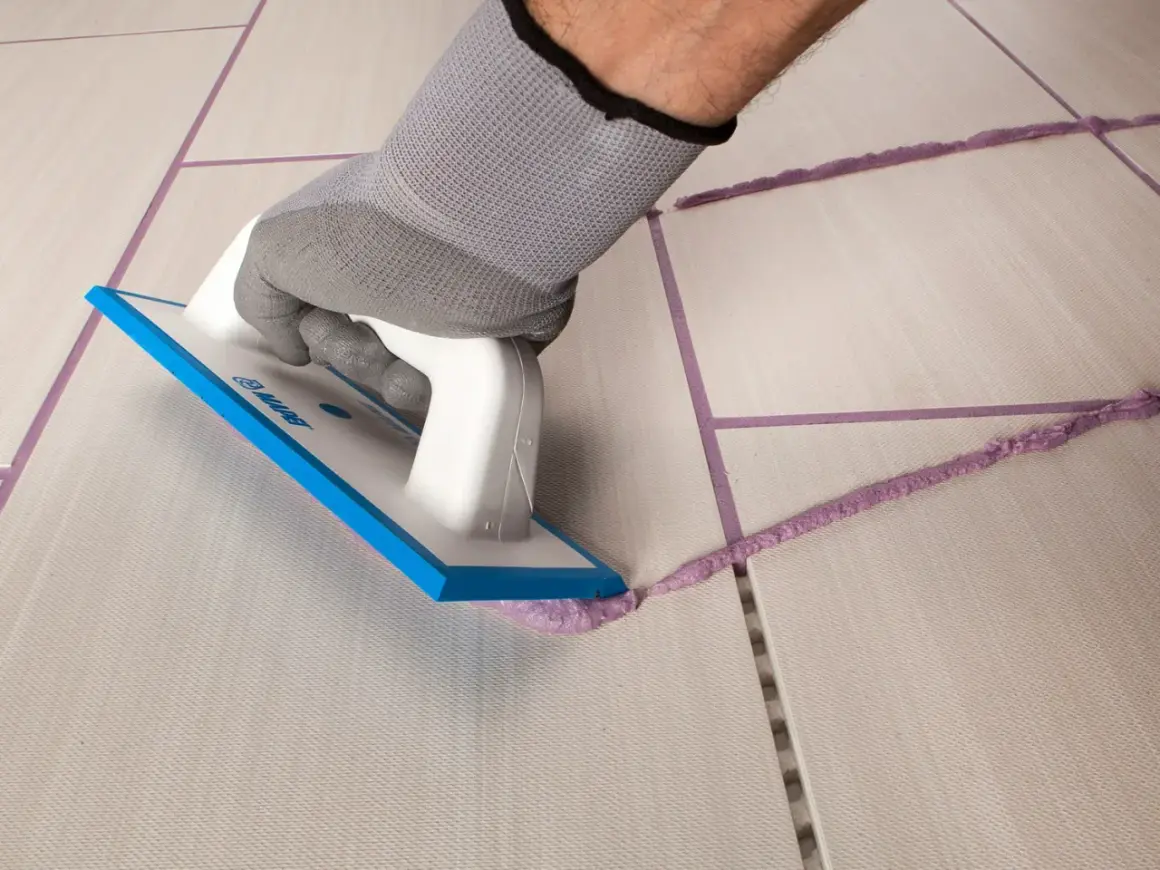 Mano con guanto grigio stende cemento e acqua senza sabbia con spatola su fughe di piastrelle.