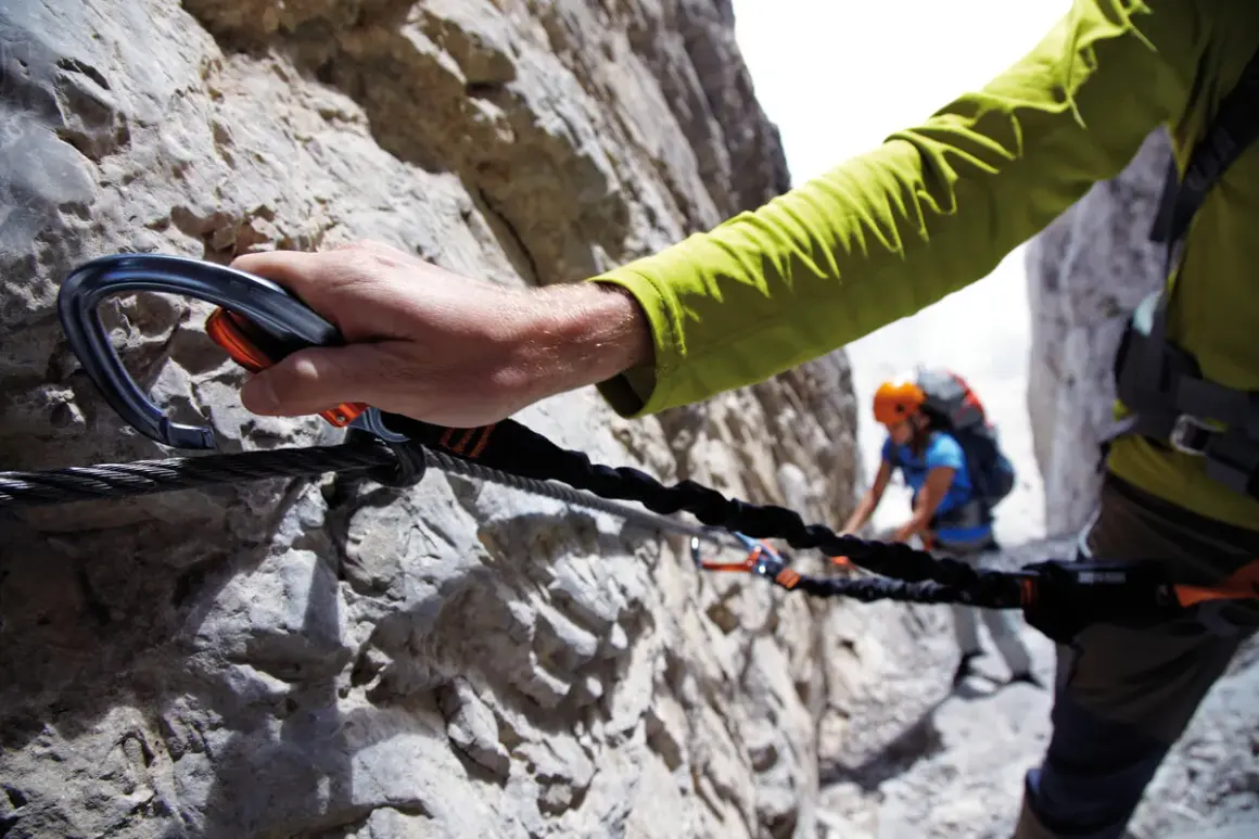 Mano agganciata a cavo d'acciaio su una parete rocciosa, un'avventura tra le ferrate Trentino con un'altra persona sullo sfondo.