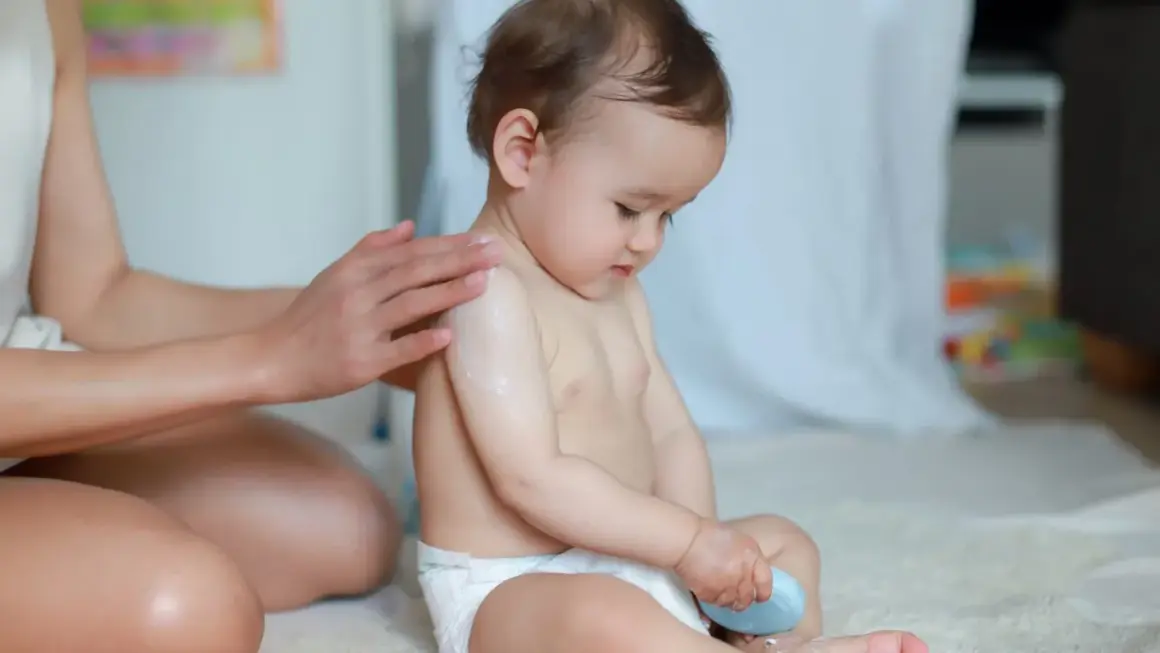 Mama smaruje kremem ramię dziecka, które siedzi w prawidłowym siadzie. Dziecko trzyma w rączce niebieską kostkę.