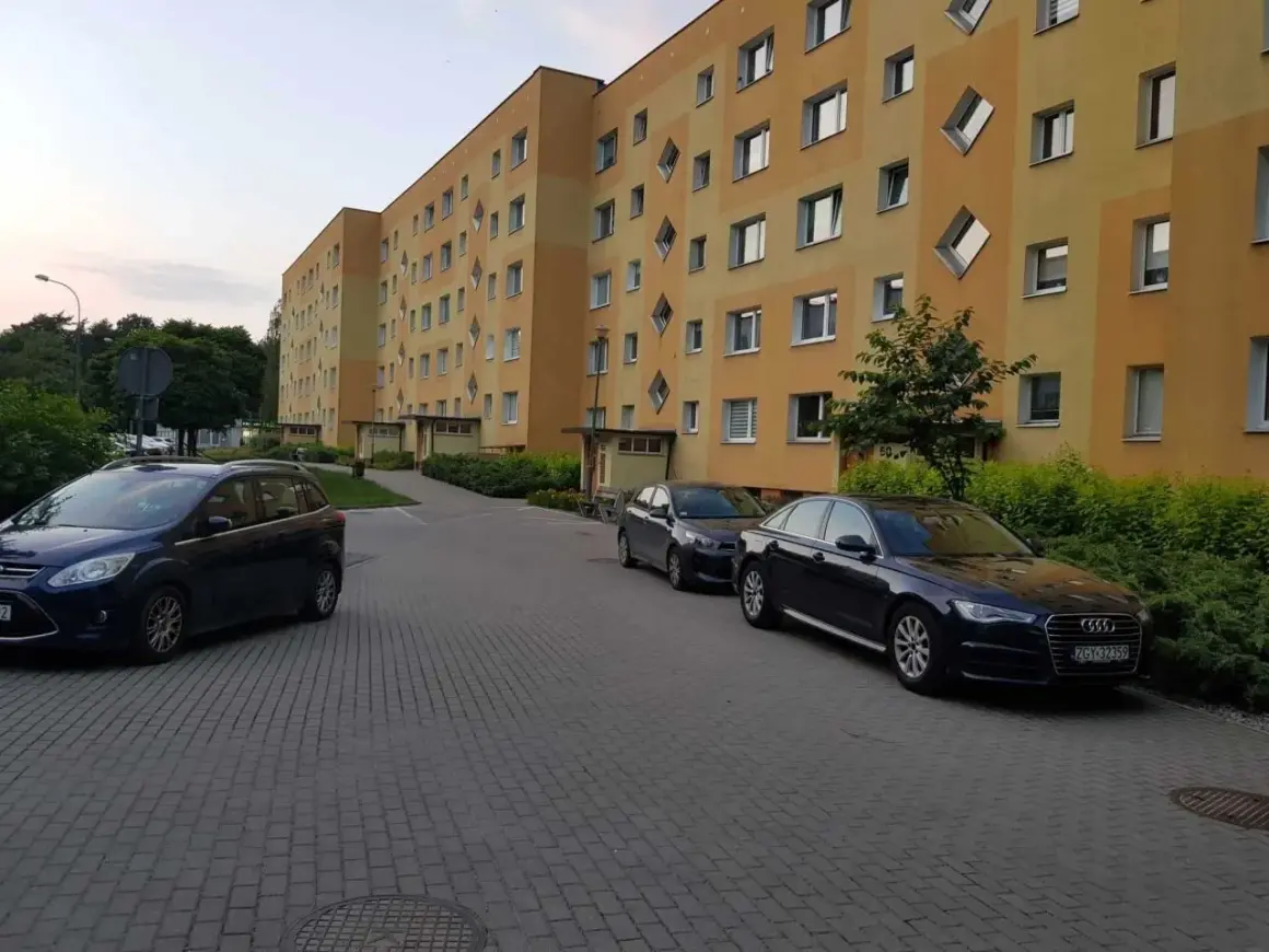 Osiedle Tysiąclecia Poznań. Jasnożółty blok z charakterystycznymi oknami w kształcie rombów, zaparkowane samochody na brukowanym podjeździe.